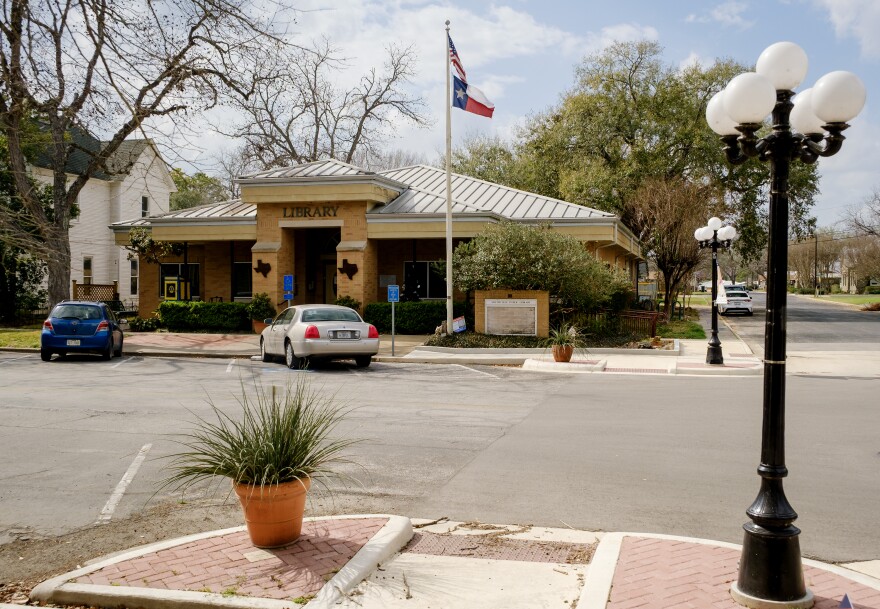 The Smithville Public Library sits across the street with an American flag hanging in front of it. In the foreground is a bush and a lamp post. 