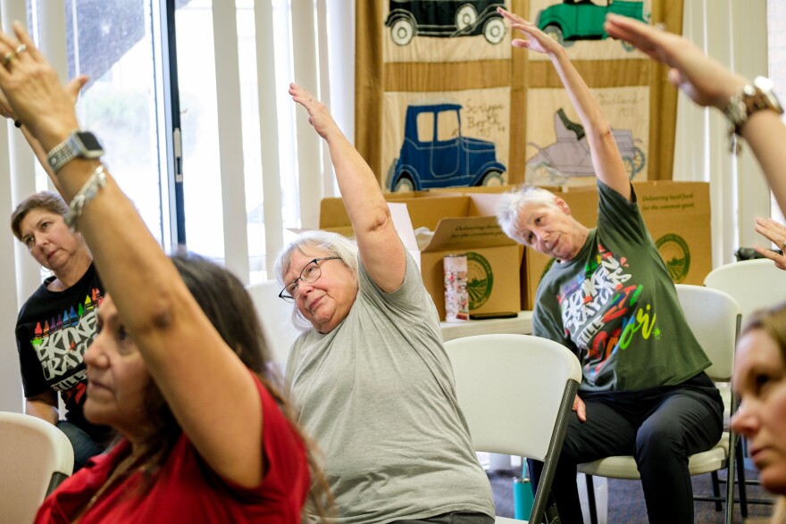 Image of a group of seated women with their left arms extended above their heads. 