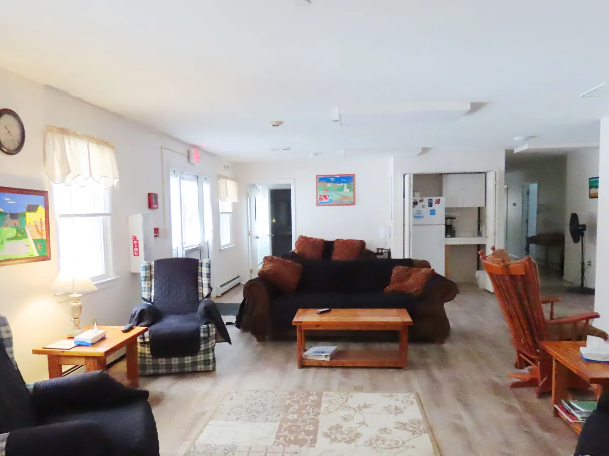 A living room with two armchairs, a dark sofa, wooden coffee tables, rocking chairs, wall art, and a view of the kitchen area. Natural light fills the space.