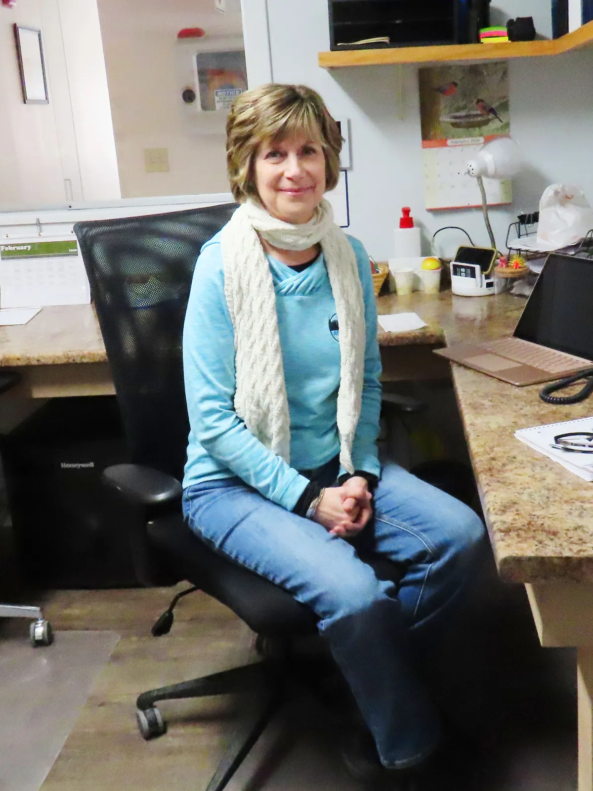 A woman is sitting at a desk in an office, wearing a light blue shirt, jeans and a white scarf, with her hands folded in her lap and a slight smile.