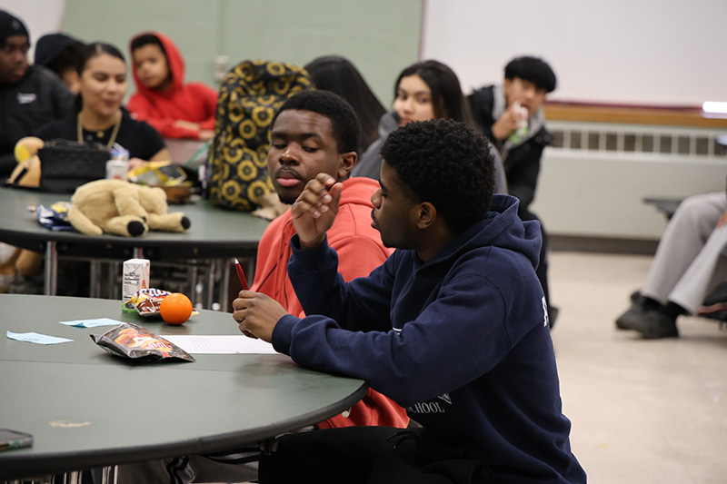 High school kids sit at tables in the cafeteria. A young man speaks.
