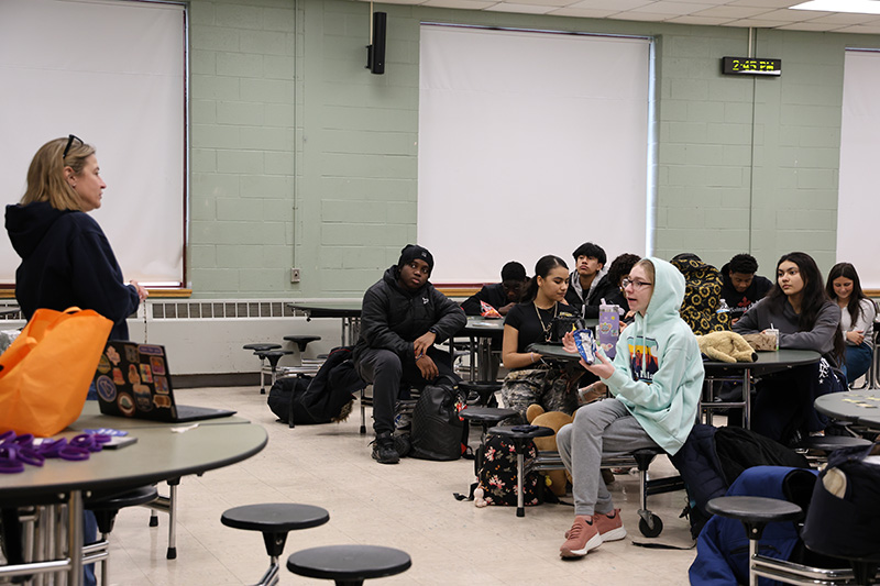 A woman in a blue sweatshirt stood in front of a large group of high school students sitting at desks.