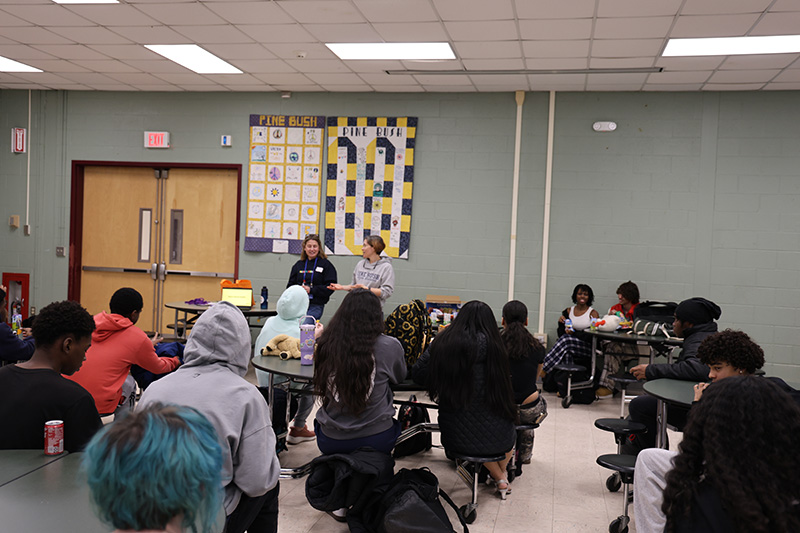 From the back of the room, a large group of high school students sit at desks discussing a woman's mental health.