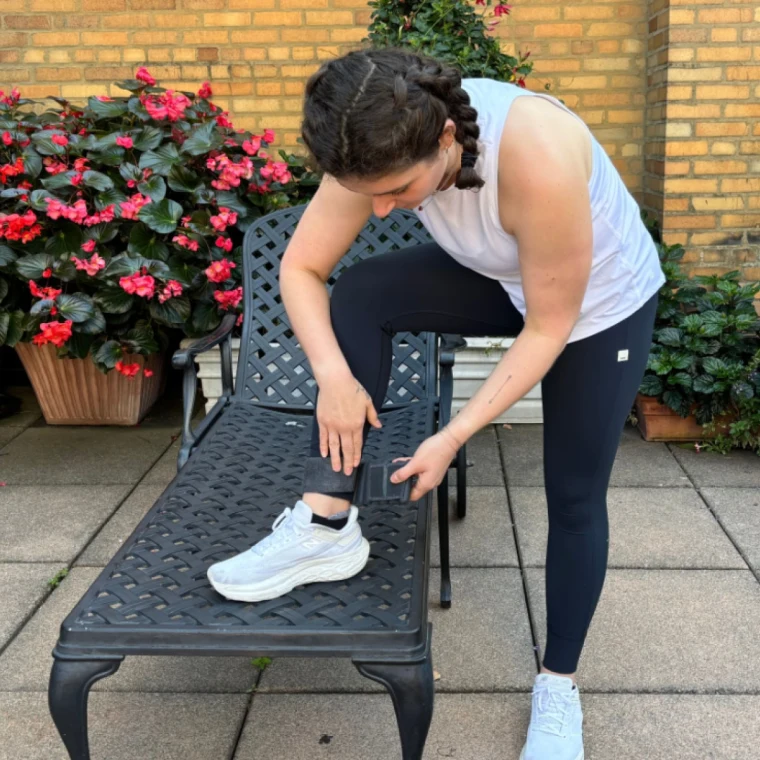 A woman stands outside on one leg on a lounge chair, leaning over to wrap a black ball bangle around her leg.