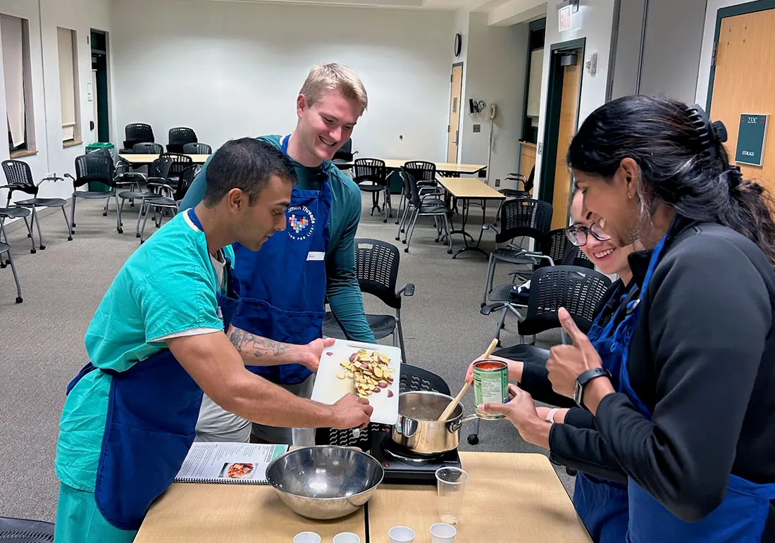 Akash Patel, wearing a University of Miami T-shirt and stethoscope, stands indoors, with tables, medical supplies, and equipment arranged behind them in a clinical training facility.