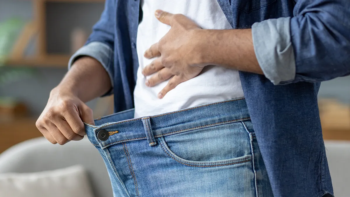 A man shows how much weight he has lost by holding the waist of his jeans, a symbol of his successful diet.