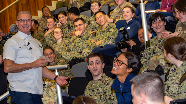 Robert Irwin in a white chef's coat stands next to a woman in a US Navy uniform, both smiling at the camera.