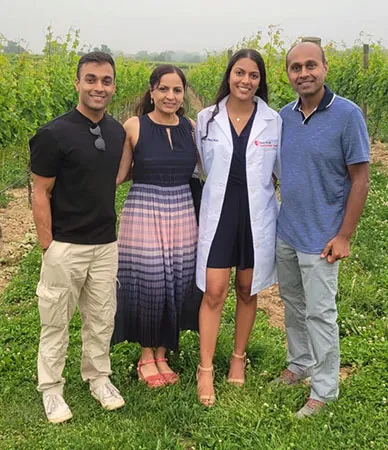 Akash Patel and family stand together outdoors in a green vineyard, posing closely with arms around each other. One person is wearing a white medical coat in a dark suit, while the others are in casual and semi-formal attire.