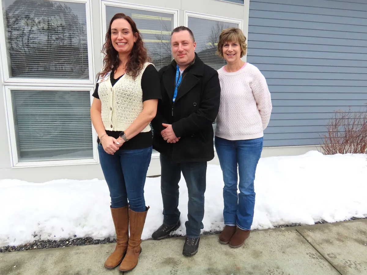 Three adults stand outside on a snowy street in front of a building with large windows and blue siding.