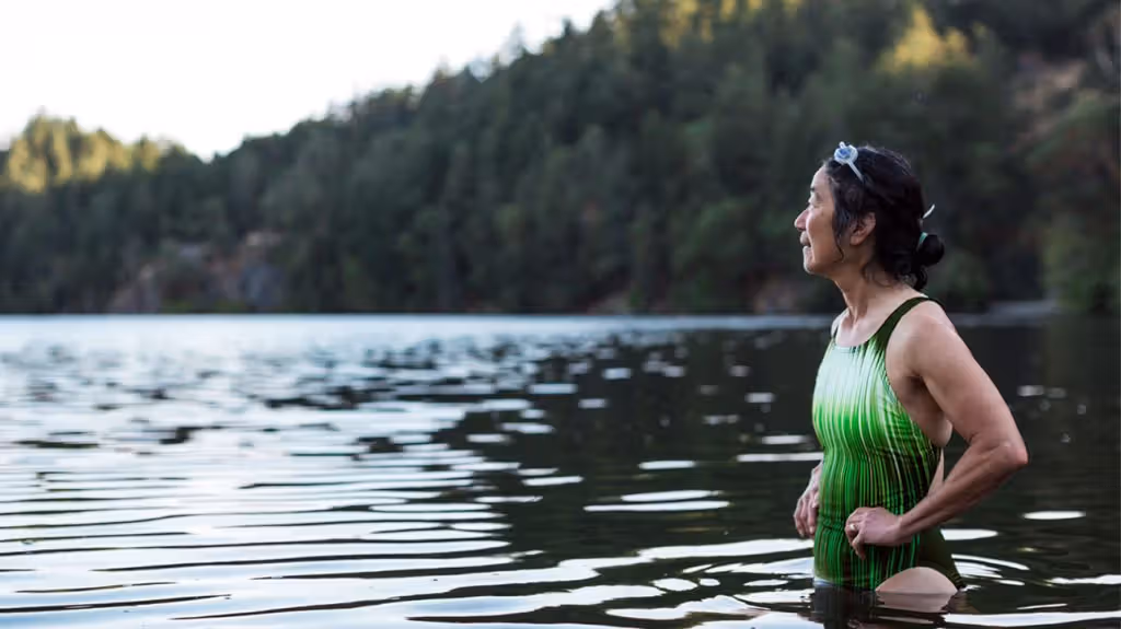 An old East Asian woman is standing in a green bathing suit by a lake