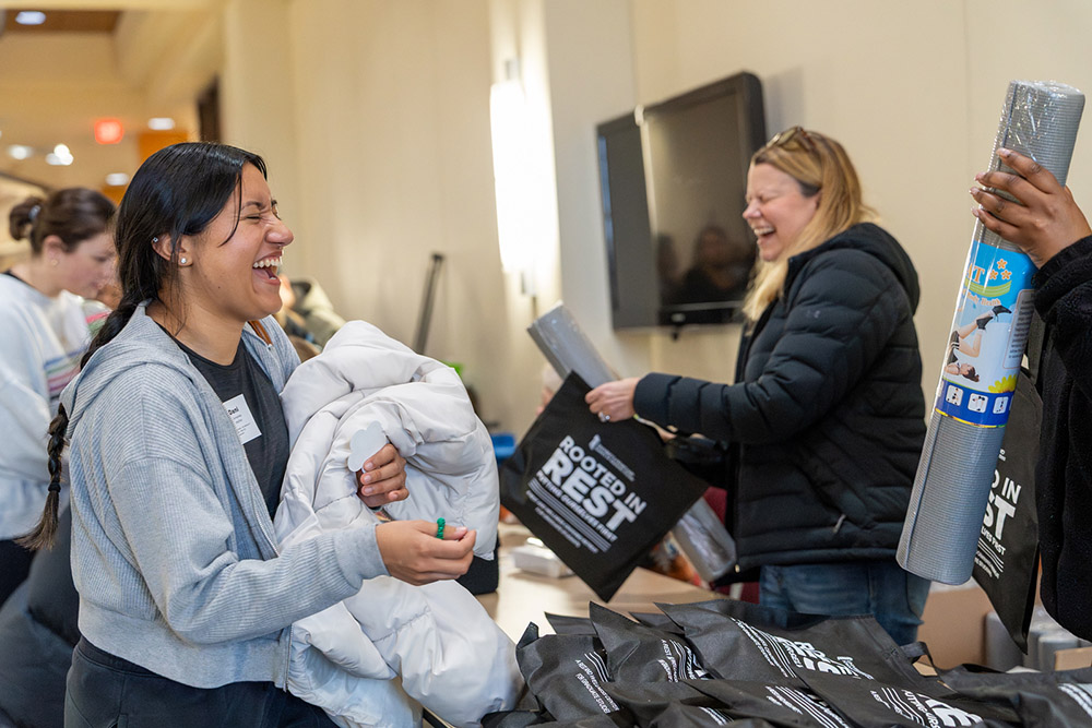 A woman smiles and holds a white jacket while interacting with others at a live event. They have yoga mats and backpacks, offering a happy atmosphere.