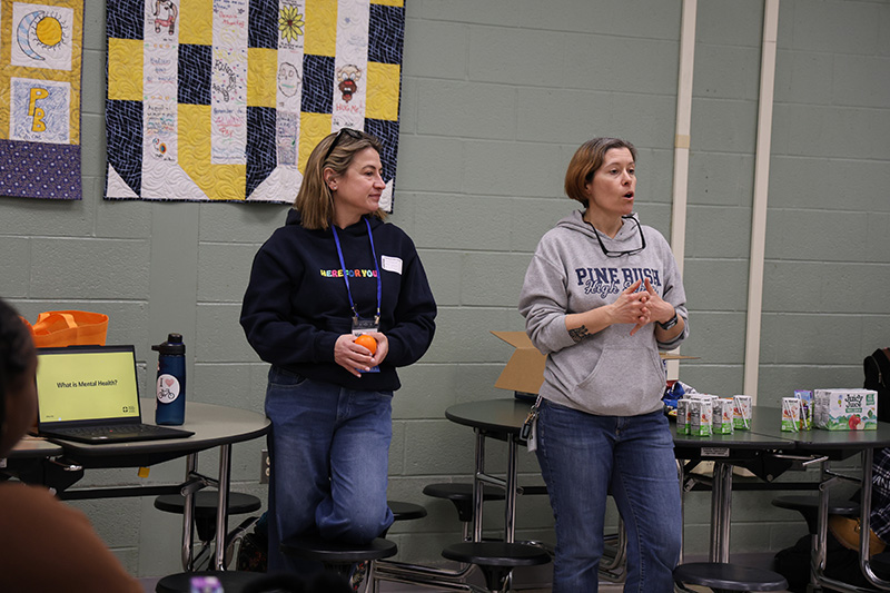 Two women stand in front of a group of high school students, talking to the children.